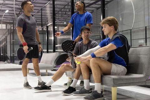 Diverse men chatting after padel match on indoor glass courts holding paddles and ball