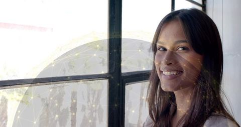 Young woman smiling by industrial window with digital network overlay, workplace portrait