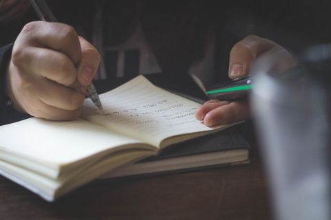 Person Writing in Notebook While Using Smartphone