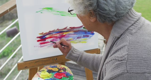 Elderly man engaging in painting a colorful abstract artwork on canvas outdoors, with paint palette close by. Ideal for illustrating creativity, senior activities, outdoor leisure, hobbyist endeavors, promoting art therapy, joyful aging, or creative expression in later life.