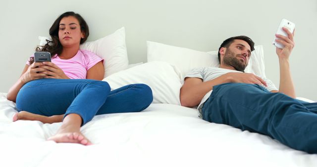 Couple lying in bed, each using their smartphones and looking distant from each other. Could be used for articles on relationship issues, technology impact on human connection, or promoting awareness about smartphone addiction and its effect on personal relationships.