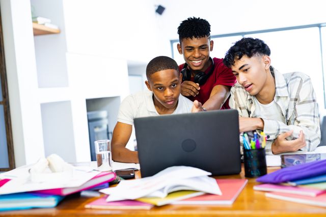 Three diverse teenage boys are gathered around a laptop at home, engaging in a collaborative activity. They appear to be studying or working on a project together, with one pointing at the screen and the others paying close attention. The scene is casual and filled with various school supplies, indicating a productive yet relaxed environment. This image is ideal for illustrating concepts related to education, teamwork, friendship, and modern youth lifestyle.