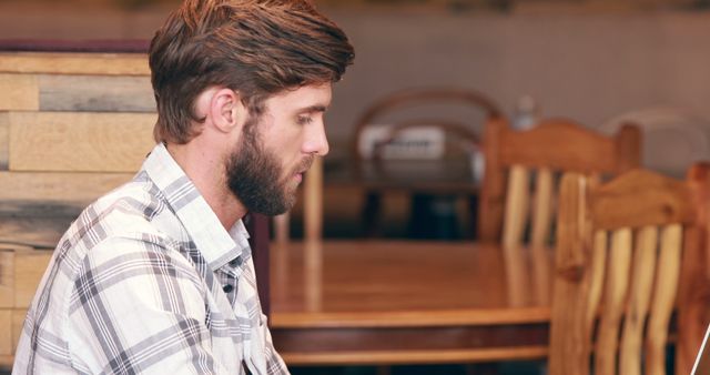 A young Caucasian man with a beard is focused on his work at a wooden table, with copy space. His casual attire and the rustic setting suggest a relaxed work environment or a creative session.