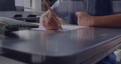 Student writing at school desk holding wooden pencil with plaid pencil case nearby