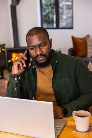 African american man working on laptop while talking on smartphone in cozy home office