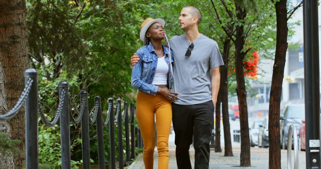 An African American woman and a Caucasian man, both young adults, are walking together on a city sidewalk, with copy space. They appear to be enjoying a leisurely stroll, indicating a romantic connection or close friendship.