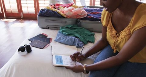 African american woman packing suitcase and writing travel checklist, planning vacation