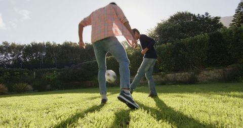 Father and son engaging in a soccer game on a lush green lawn, backlit by the sun, creating a heartwarming family scene. Perfect for illustrating family bonding moments and the joy of outdoor leisure in residential areas. Suitable for advertisements focusing on family, sports, lifestyle, or outdoor products.