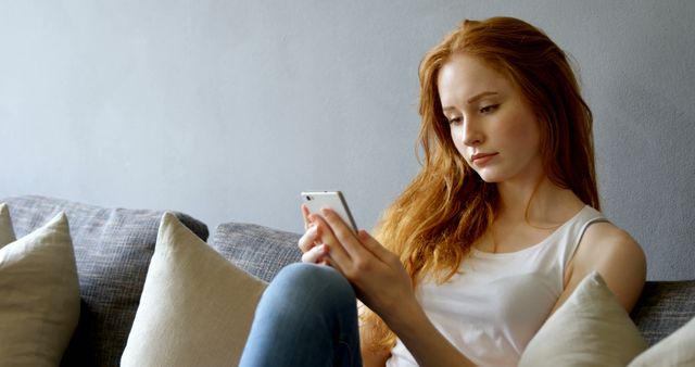 A young redhead woman sits on a comfortable couch, holding and texting on her smartphone. Her casual attire and thoughtful expression suggest she is engaged in a serious conversation or reading something important. Ideal for use in articles or promotions related to social media, technology usage, communication, home lifestyle, or youth culture.