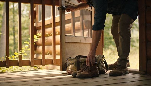 Traveler reaching for boots on rustic cabin porch with duffel bag and sunlit forest