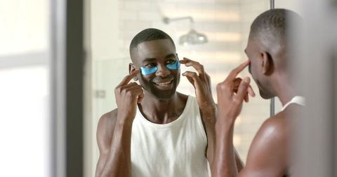 African american man applying blue under-eye gel patches at bathroom mirror for skincare routine