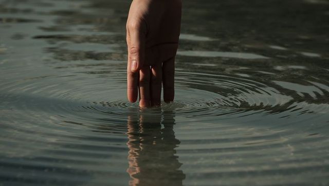 Human hand touching calm pond water creating concentric ripples and soft reflection