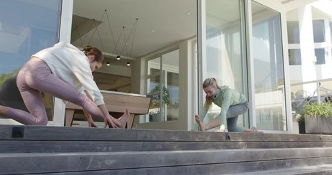 Two women stretching on wooden deck steps by glass doors outdoor morning workout