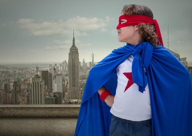 Digital composition of superhero kid in blue cape and red eye mask standing with cityscape in background