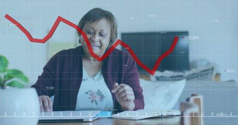 Mature woman managing finances at home with laptop and red stock market chart overlay