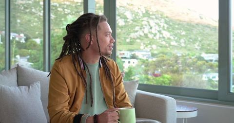 Mid-adult man with dreadlocks relaxing on sofa holding mug while gazing out large windows