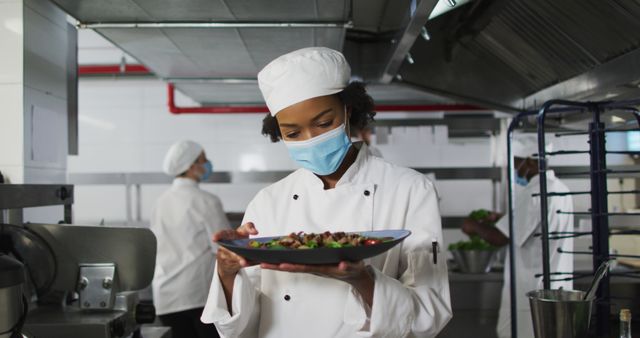 Portrait of african american female chef wearing face mask presenting dish. Health and hygiene in restaurant kitchen during coronavirus covid 19 pandemic.