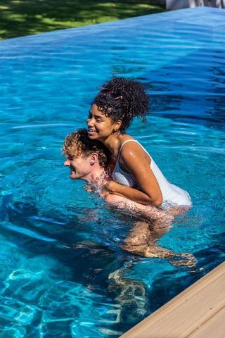 Laughing diverse couple playing piggyback in sunlit pool with crystal-clear blue reflections