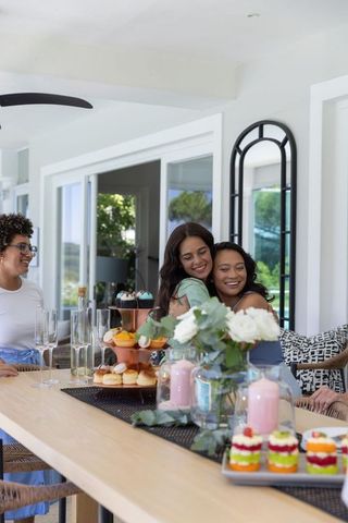 Diverse women hugging at bright brunch table with desserts, champagne, floral centerpiece