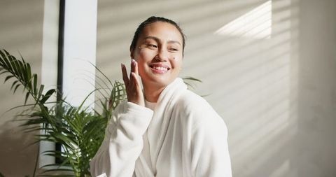 Asian woman applying face cream in sunlit robe by window with plants and blinds shadows