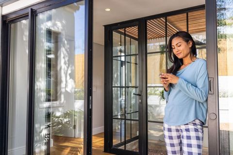 Asian woman standing at glass doors holding smartphone, wearing pajamas, morning light