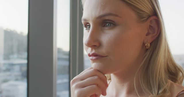 Young blonde woman looking through window thoughtfully, hand on chin. The image captures a moment of contemplation and introspection, with details like earrings adding a personal touch. Ideal for use in content related to introspection, decision-making, mindfulness, beauty, lifestyle, or even mental health.
