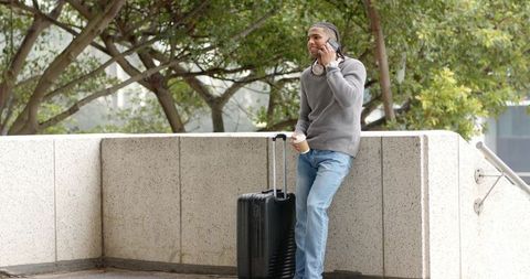 African American man leaning on concrete wall calling on phone with suitcase and coffee