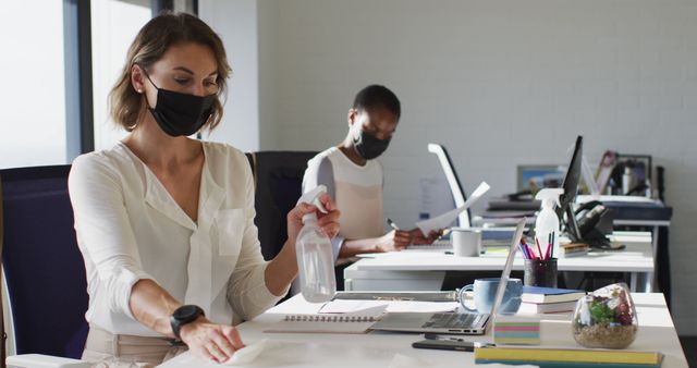 Image depicts professional office workers wearing face masks and cleaning their desktops. They are practicing good hygiene and following COVID-19 safety protocols. This can be used in articles related to workplace hygiene, pandemic precautions, office environments, safety measures, and teamwork.