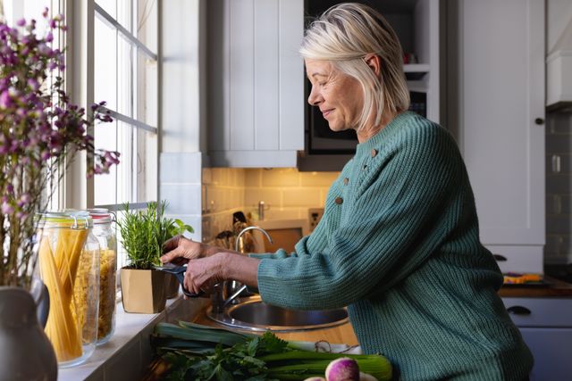 Smiling mature caucasian woman standing by kitchen window cutting herbs, copy space. Domestic life, living alone and senior lifestyle concept.