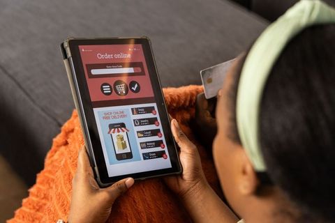 African American woman holding credit card browsing online store on tablet on cozy couch