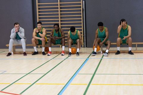 Diverse male basketball team relaxing on bench at indoor basketball court