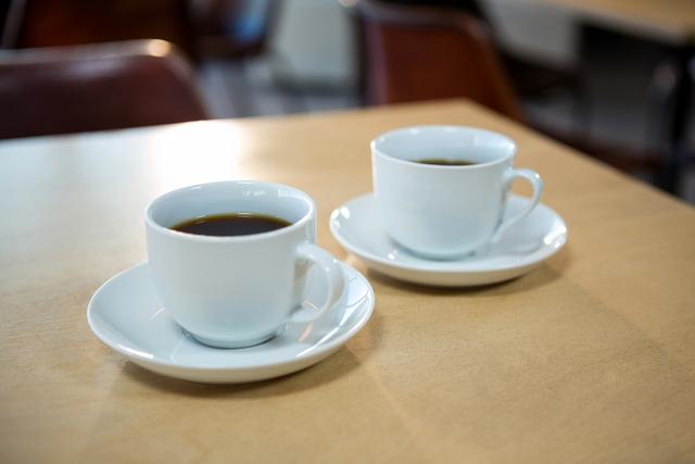 Two white cups of black coffee on a wooden table in a cafeteria. Ideal for illustrating concepts of relaxation, coffee breaks, morning routines, or cafe settings. Suitable for use in blogs, advertisements, or social media posts related to coffee culture, cafes, or beverages.