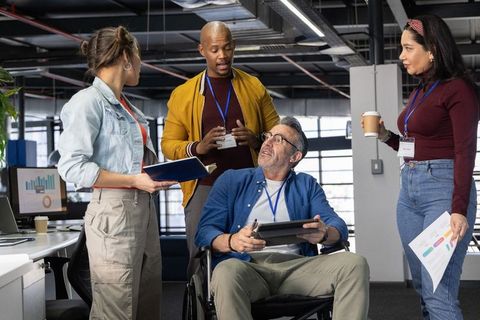 Diverse team collaborating around colleague in wheelchair in modern open-plan office