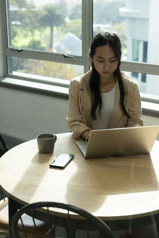 Asian woman wearing beige blazer working on laptop at round table in sunlit modern office