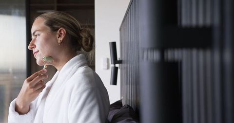 Woman using jade facial roller in white robe on bed for morning skincare routine