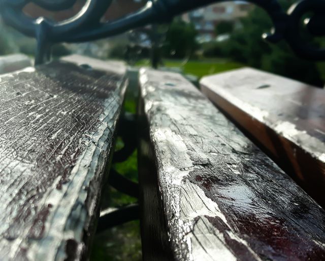 Wooden garden bench seen close-up under direct sunlight in garden or park reveals its aged and weathered texture with hint of metal armrest. Ideal for resources related to outdoor furnishings, rustic wares, garden decoration, or natural aging. Can be used in blogs and articles focusing on relaxing outdoor environments or urban parks.