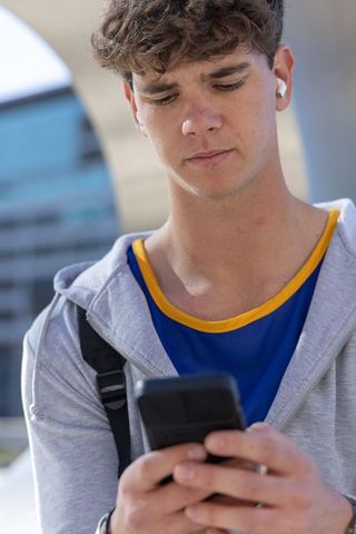 Teen checking smartphone in urban plaza wearing hoodie, wireless earbuds and backpack