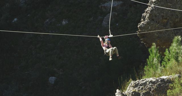 Person wearing safety gear ziplining through a forested area with rocky surroundings. Perfect for promoting outdoor adventure activities, tourism brochures, and extreme sports advertisements.