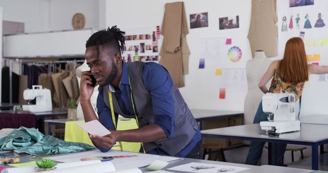 Young African American man talks on the phone in a busy design studio. He coordinates tasks while a young Caucasian woman works on a dress nearby.