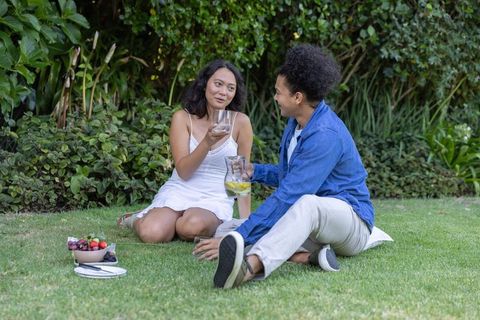 Multicultural couple sitting on lawn pouring lemon water and chatting at casual backyard picnic