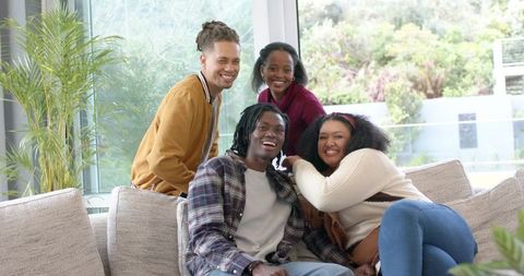 Smiling diverse friends relaxing together on modern sunlit sectional sofa with plants