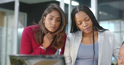Diverse female coworkers collaborating over laptop in modern office reviewing report