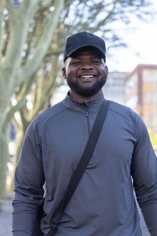 Smiling man in black cap and zip pullover walking with crossbody bag in city