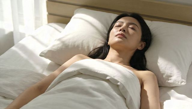Sunlit asian woman resting on minimalist white bed, serene morning sleep and wellness