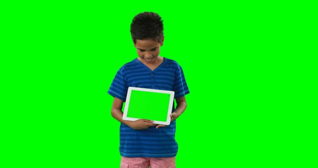 A young boy stands against a green screen holding a tablet with a green screen, with copy space. His casual attire and the digital device suggest a context of modern technology use among children.