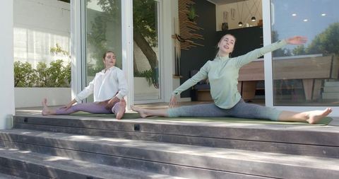 Two women stretching in full split on sunny wooden patio practicing outdoor yoga