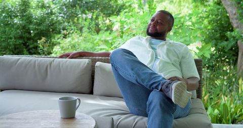 African american man relaxing on outdoor wicker sofa with coffee cup and cushions