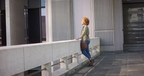 Young woman leaning on concrete railing on urban terrace carrying brown leather bag