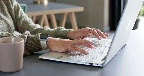 Hands typing on white laptop wearing knit sweater and smartwatch, cozy home workspace