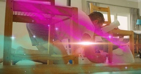 Kneeling man reaching under wooden table for toy ball with cosmic nebula light overlay